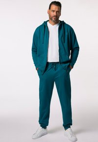 Teal zip-up hoodie med framfickor och matchande joggers, kombinerat med vita sneakers. Tillverkad av mjukt, slätt tyg med en avslappnad passform.