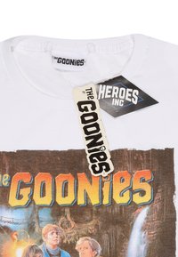 Bílá tričko s grafikou "Goonies" a barevným textem a postavami, doplněné o tagy "Heroes Inc" a "Goonies."