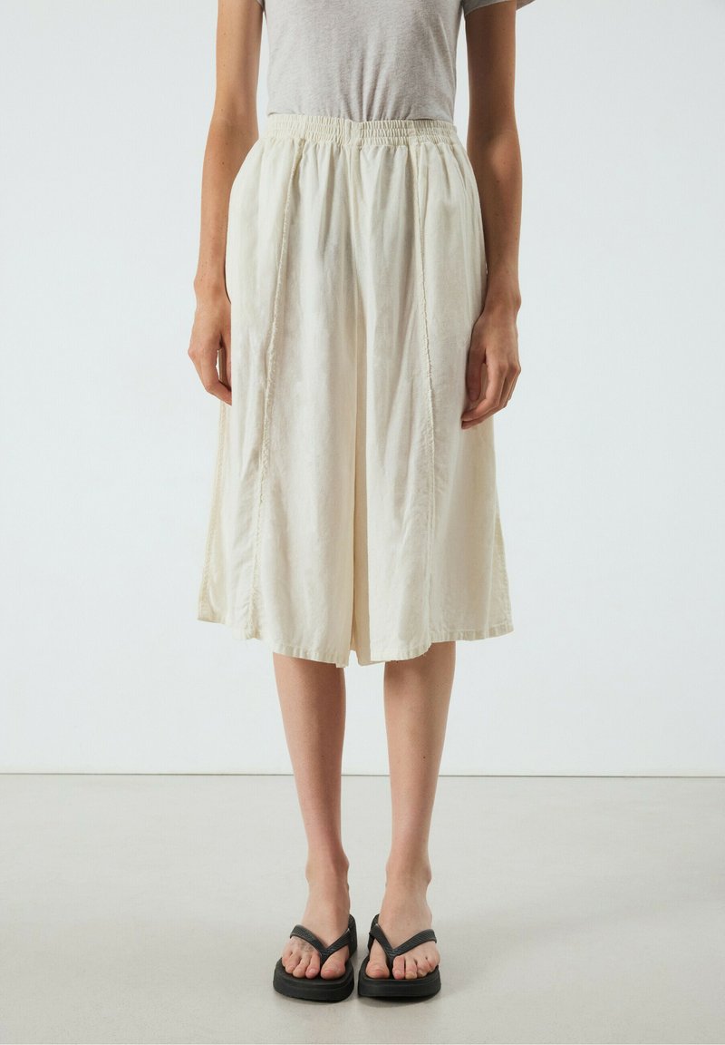 Femme portant une jupe ample couleur crème longueur genou, une chemise à manches courtes gris clair et des tongs à plateforme noires, debout sur un sol uni.