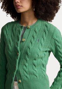 Grön kabelstickad cardigan i mjukt material, med rund halsringning, tre knappar och en liten lila logotyp på bröstet.