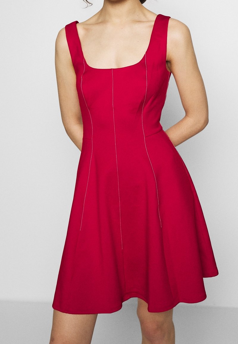 Femme portant une robe rouge sans manches, longueur genoux, avec un col carré et des coutures verticales visibles.