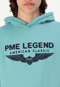 Turkusowa bluza z kapturem z grafiką z przodu z tekstem "PME LEGEND" i "AMERICAN CLASSIC", akcentowana skrzydlatym emblematem i skomplikowanym haftem.