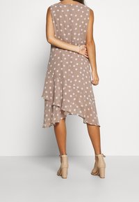 Braunes Kleid mit Polka-Dots und mehrlagigem Saum. Ärmelloses Design mit geknöpften Rücken. Getragen mit beigen Sandalen mit Absatz.