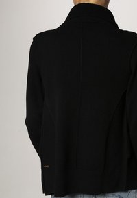 Svart stickad cardigan med hög krage, ribbad textur och detalj i sidosen. Accenterad med en liten guldfärgad logotyp på nedre vänstra sidan.