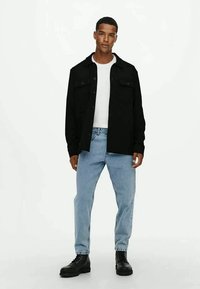 Camisa preta de botão com dois bolsos, usada sobre uma t-shirt branca; emparelhada com jeans azul claro e botas pretas com atacadores.