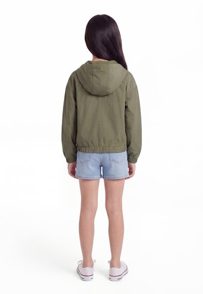 Child with long dark hair wearing an olive green hooded jacket, light denim shorts, and white sneakers, standing facing away.