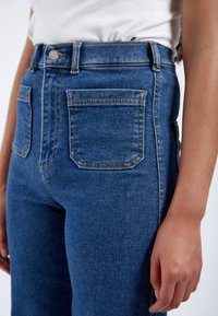 Jeans de tiro alto en azul, hechos de denim, con cinco bolsillos, costuras claras y cierre frontal con botón. Textura suave, forma ajustada.