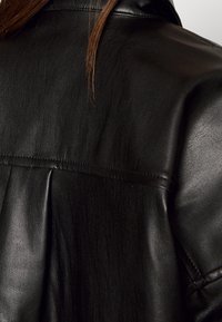 Svart läderjacka med en slät yta och subtil glans, med en plisserad detalj på baksidan och kontrasterande sömmar längs kanterna.