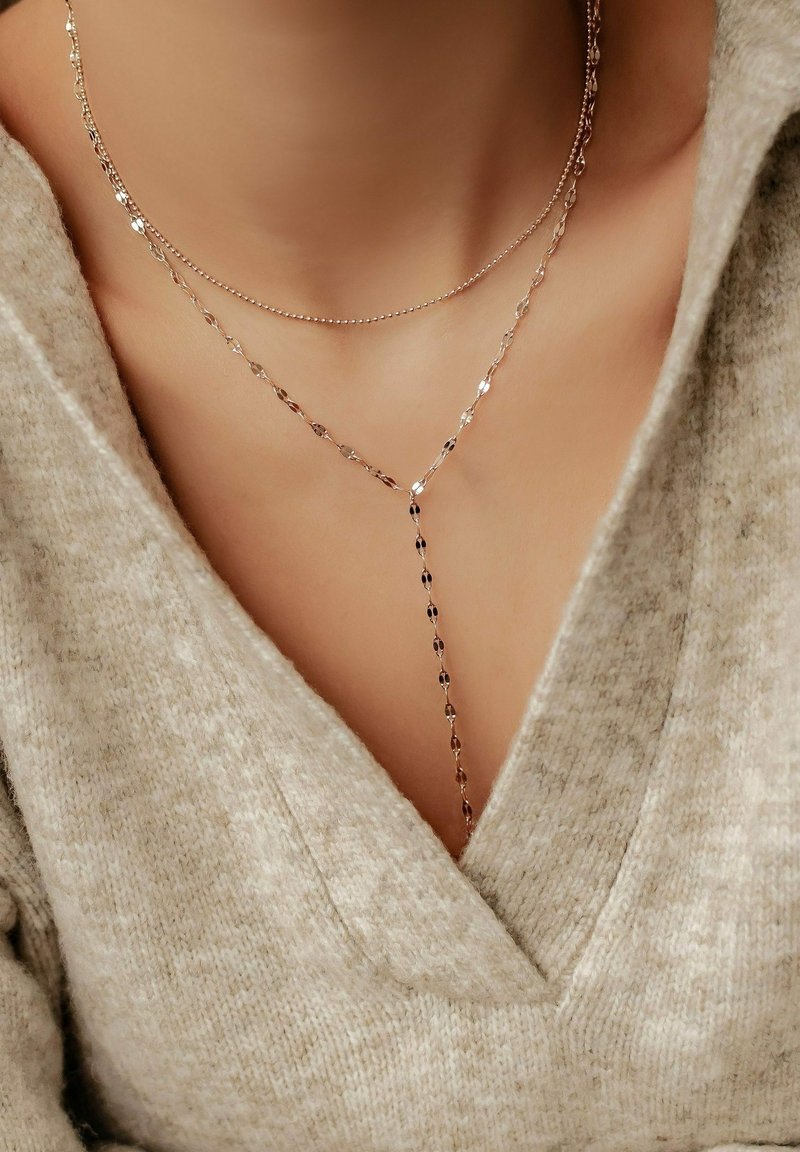 Deux colliers délicats en chaîne d’or sur un cou, l’un court et l’autre plus long avec une goutte, portés avec un pull en maille beige à col en V.