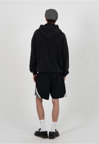 Sweat à capuche noir surdimensionné avec une capuche, associé à un short noir avec des accents gris sur les côtés. Porté avec des chaussettes blanches et des chaussures noires.