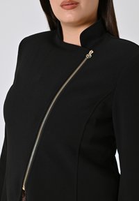 Zwarte jas van gladde stof, met een hoge kraag, asymmetrische gouden ritsdetails aan de voorkant en een getailleerde pasvorm.
