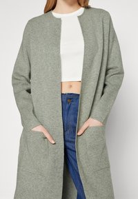 Salvgrön lång cardigan i mjukt tyg med rund hals, öppen framsida och två sidfickor. Bärs över en vit topp och jeans.