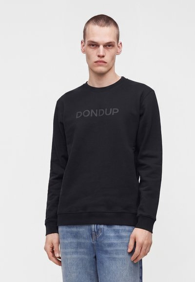Млад мъж с къса коса, носещ черна блуза с лого "DONDUP" и светлосини дънки, стои пред обикновен бял фон.