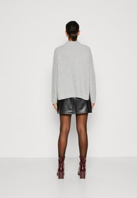 Žena stojící čelem k nám, oblečená v šedém pleteném svetru, černé kožené sukni, průhledných punčochách a vínově červených kotníkových botách.