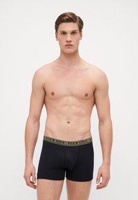 Schwarze Baumwoll-Boxershorts mit einem tanfarbenen Gummibund, der den Text "PIER ONE" in einem wiederholenden Muster zeigt. Glatte Textur, körpernahe Passform.