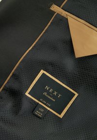 Material negru cu un finisaj texturat, având un accent de culoare auriu. Include un etichetă pentru "NEXT Occasion" și un marcaj de mărime "40R."