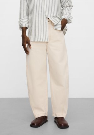 Uomo che indossa pantaloni beige a gamba larga, scarpe marroni e una camicia a righe beige e bianca con un bottone, con una mano in tasca.