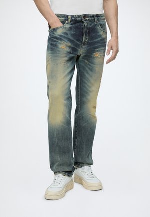 Mann trägt ausgewaschene blaue Jeans mit abgenutzten Stellen, weiße Sneaker und ein weißes Hemd. Eine Hand ist in der Tasche, steht aufrecht.