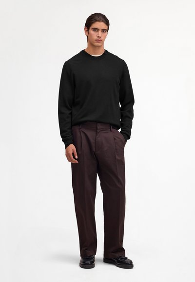 Ung mand iført en sort langærmet skjorte, mørkebrune bukser med vide ben og sorte lædersko, stående på en hvid baggrund.