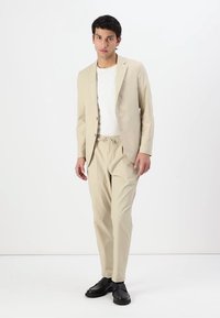 Beige oblek s upravenou bundou a kalhotami s elastickým pasem. Nosič má na sobě bílé tričko. Doplněno o černé boty. Minimalistický design.