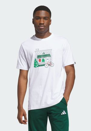 Młody mężczyzna w białej koszulce z zielonym motywem sklepu, choinki i roweru oraz ciemnozielonych spodniach Adidas z logo.