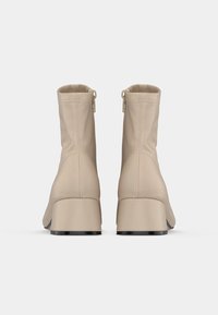 O pereche de cizme bej până la glezne, cu tocuri pătrate și fermoare la spate, prezentate din spate pe un fundal alb.