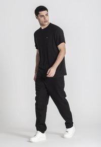 Čierne, rebrované tričko s krátkym rukávom v kombinácii s čiernymi nohavicami. Obuvou sú biele tenisky. Neutrálny pozadí zdôrazňuje detaily oblečenia.