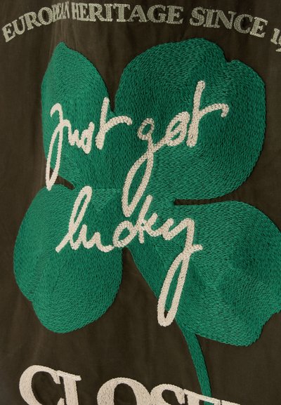 Trèfle à quatre feuilles vert brodé avec texte blanc cousu "just get lucky" sur tissu foncé et mots partiels "European Heritage Since" visibles.