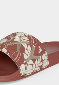 Une sandale marron à enfiler avec un motif floral blanc et des lettres beiges en relief sur la sangle.