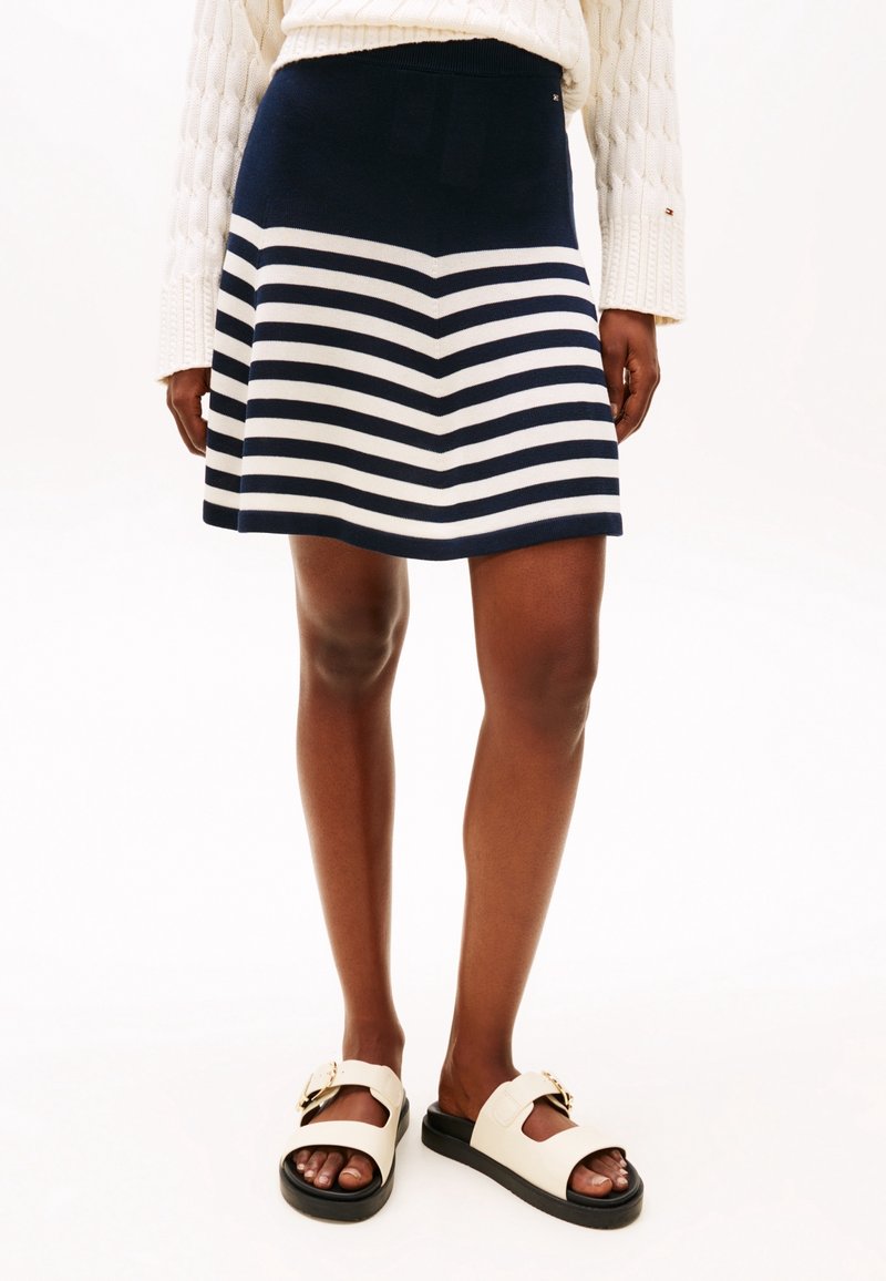 Pletená sukně s námořnickými a bílými vodorovnými pruhy, s přiléhavým pasem a rozšířeným tvarem; doplněná bílými skluznými sandály.