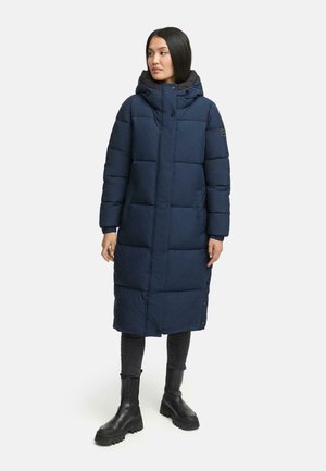 Marineblå pufferjakke med hætte, der har et kviltet design, trykknapper og en knælængde. Bæres over mørke jeans og kraftige støvler.