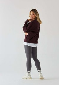 Burgundowa bluza nałożona na białą koszulę, w połączeniu z dopasowanymi szarymi legginsami i białymi sneakersami z czarnymi akcentami.