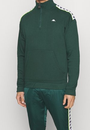 Homme portant un sweat-shirt à fermeture éclair quart vert foncé et un pantalon de jogging assorti avec des bandes de logo blanches et noires sur les épaules et les côtés du pantalon.