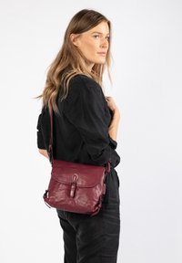 Bordeaux leren crossbody tas met een klepopening, verstelbare schouderband en subtiele stiksels. Gladde textuur met metalen accenten.
