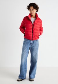 Rode pufferjack met een rits aan de voorkant, geribde manchetten en een rechte kraag; gecombineerd met lichtblauwe wijde jeans en zwartgestreepte schoenen.