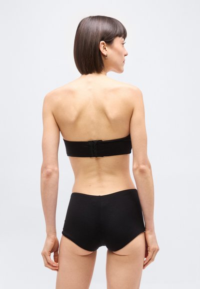 Γυναίκα με κοντά καστανά μαλλιά, που φαίνεται από πίσω, φοράει μαύρο στράπλες σουτιέν και μαύρο εσώρουχο με ψηλή μέση, μπροστά σε απλό φόντο.