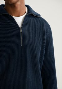 Man in een marineblauw ribgebreid trui met een gedeeltelijk ritskraag over een witte ronde hals t-shirt, weergegeven van de nek tot het midden van de torso.
