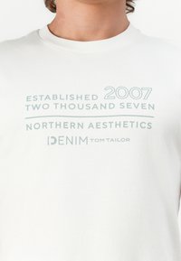 Camiseta de algodón blanca con texto impreso en azul claro: "ESTABLECIDO 2007", "DOS MIL SIETE", "ESTÉTICAS NORTEAMERICANAS" y "DENIM TOM TAILOR".