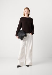 Maglione marrone a coste, pantaloni bianchi larghi e una pochette nera a pieghe. Modella che indossa scarpe nere a punta, in piedi contro uno sfondo neutro.