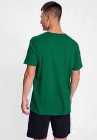 Groene katoenen T-shirt met korte mouwen, ronde hals, relaxte pasvorm. Zichtbare stikseldetails, gedragen met marineblauwe shorts. Achteraanzicht.