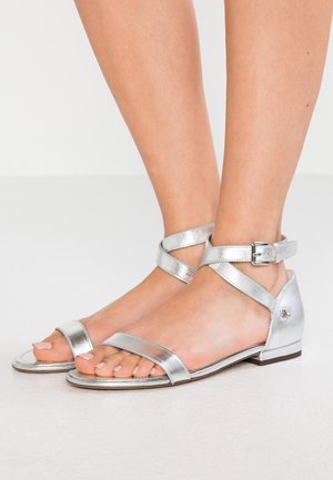 Srebrne metaliczne sandały z krzyżującymi się paskami na kostce, otwartym noskiem, płaską podeszwą i małym dekoracyjnym elementem na obcasie.