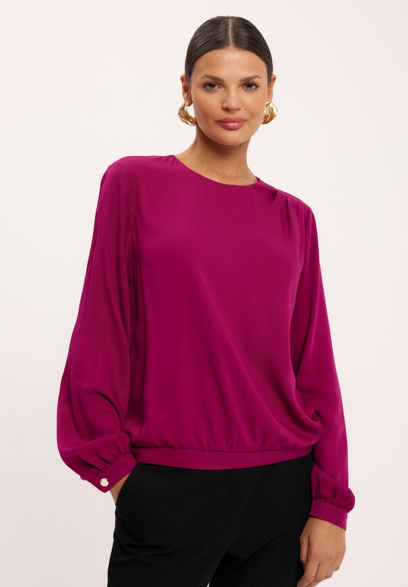 Magenta bluzka z okrągłym dekoltem, długimi bufiastymi rękawami i zebranym dołem. Miękka tkanina z minimalnymi detalami i luźnym krojem.