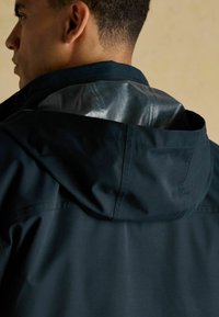 Tmavomodrá bunda s kapucňou so hladkým, lesklým materiálom na hornej časti chrbta. Tkanina má elegantný povrch s viditeľnými detailmi šitia.