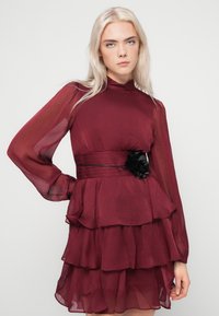 Νεαρή γυναίκα με μακριά ξανθά μαλλιά, φορώντας ένα στρωματοποιημένο μπορντό φόρεμα με διαφανείς μακριές μανίκες και μια μαύρη ζώνη με λουλούδια, που στέκεται μπροστά σε απλό φόντο.