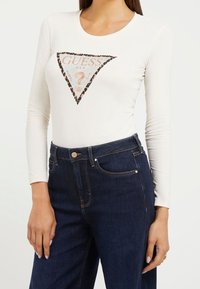 Camicia bianca a maniche lunghe con un design stampato triangolare che presenta dettagli in stampa leopardata, testo del logo, abbinata a jeans in denim scuro.