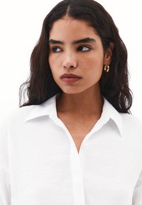 Camicia bianca con colletto strutturato e leggero texture, abbinata a orecchini a cerchio dorati e incorniciata da capelli scuri e mossi.