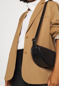 Brauner Blazer mit zwei Knöpfen, kombiniert mit einem weißen Rollkragenpullover. Eine schwarze Leder-Schultertasche mit gebogener Form lehnt am Körper.