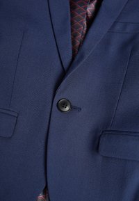 Blazer blu navy con texture liscia, dotato di un bottone nero, tre tasche e una fodera con motivo geometrico rosso in vista.