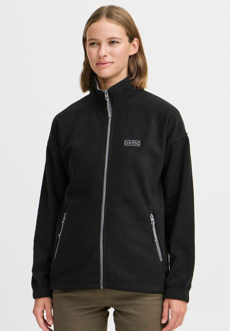 Musternahk fleece-jakk musta, millel on kõrge kaelus, täispikk tõmblukk ja kaks tõmblukuga taskut. Esiküljel on peen "oxmo" logo valges värvuses.