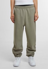 Pantaloni de trening ajustați din bumbac verde prașmuit, cu un talie elastică și glezne strâmte. Purtați cu adidași albaștri.